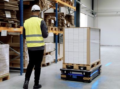 Warehouse worker in a safety vest walking beside an autonomous pallet robot